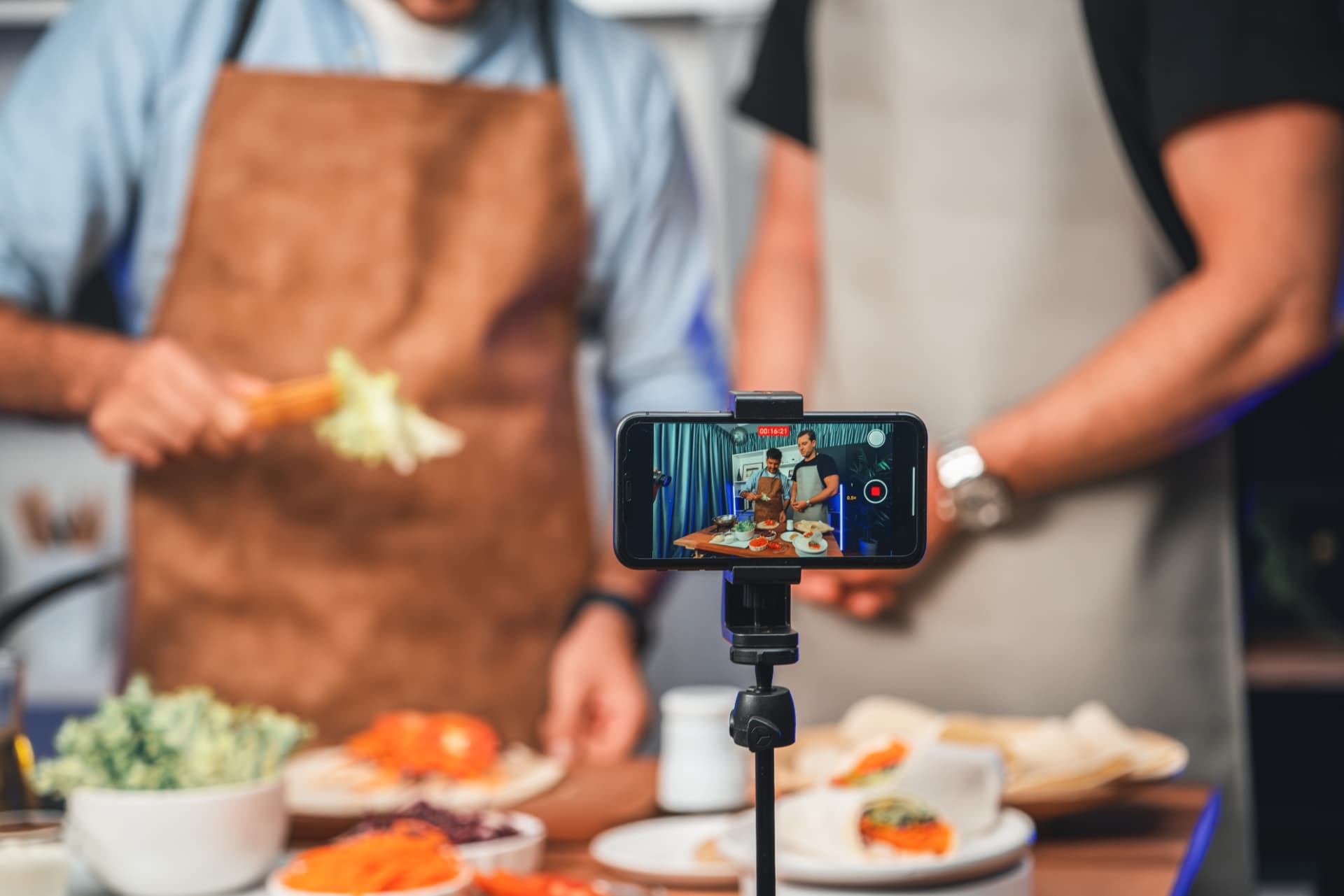 Smartphone em tripé gravando dois chefs preparando pratos em uma cozinha profissional