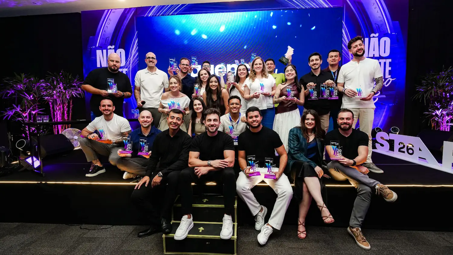 Grupo de profissionais de agências de marketing premiados em palco durante evento do setor no Brasil, segurando troféus em frente a painel iluminado em azul