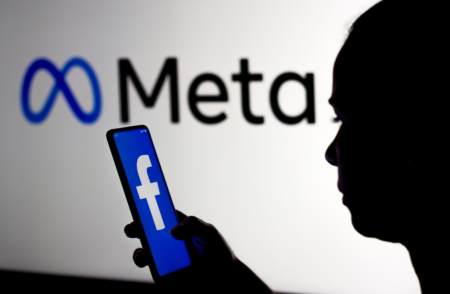 Silhueta de pessoa segurando smartphone com o logo do Facebook em frente a painel iluminado com a marca Meta