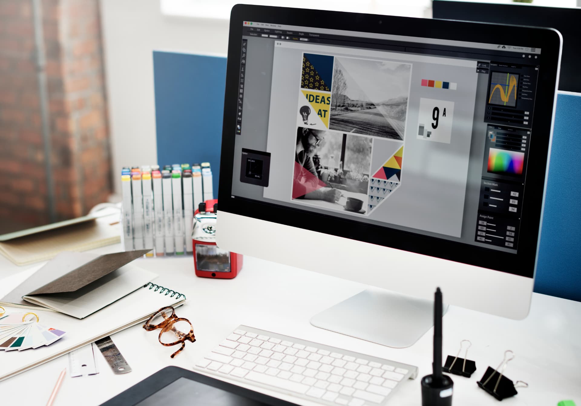 Mesa de trabalho criativa com monitor exibindo projeto de design, marcadores e caderno, ilustrando a rotina de uma agência de marketing