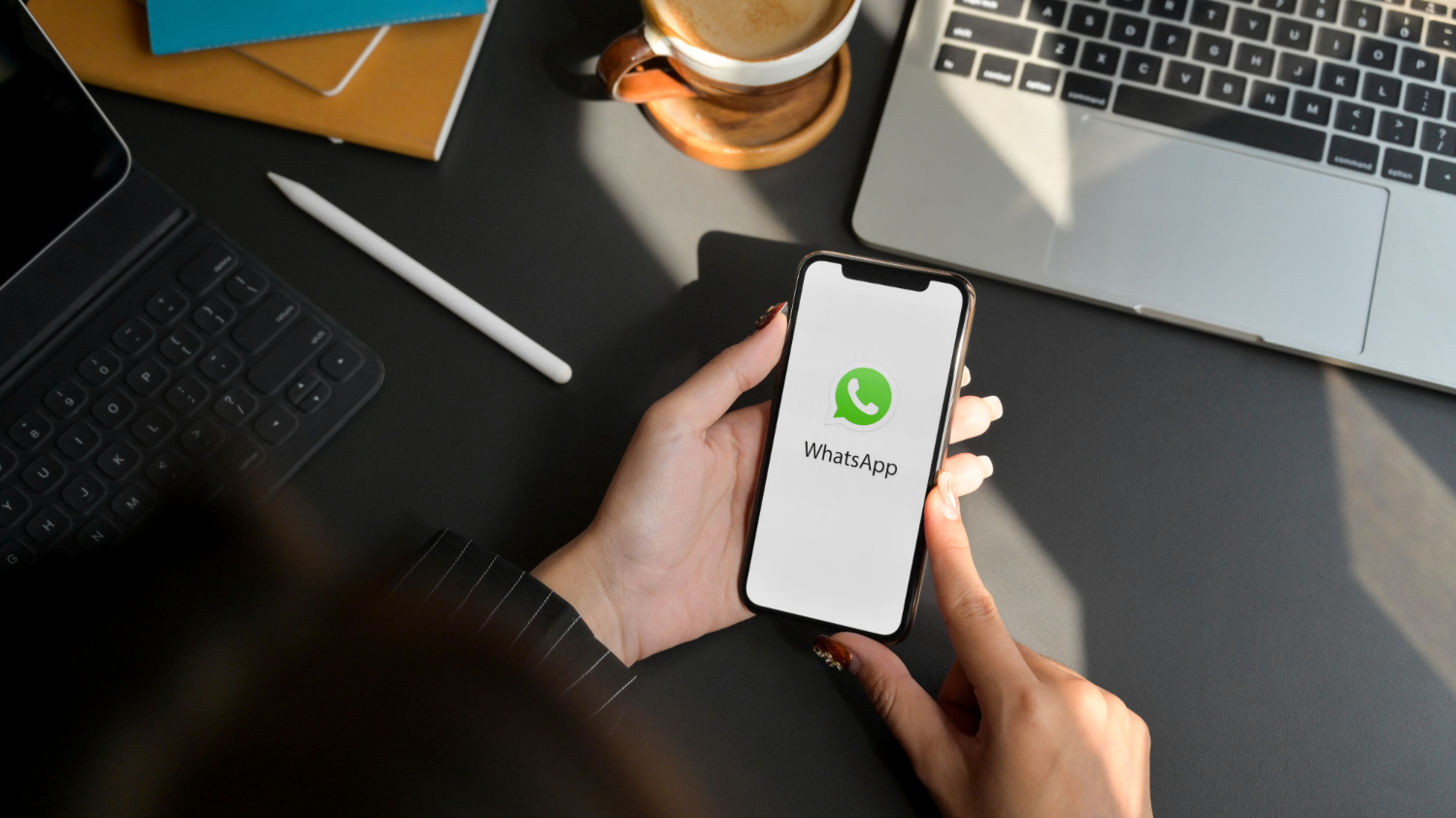 Mãos segurando smartphone com a tela do WhatsApp aberta sobre mesa de escritório com notebook e xícara de café
