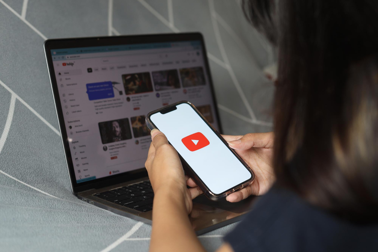 Pessoa segurando smartphone com o logo do YouTube em frente a notebook aberto na página inicial da plataforma