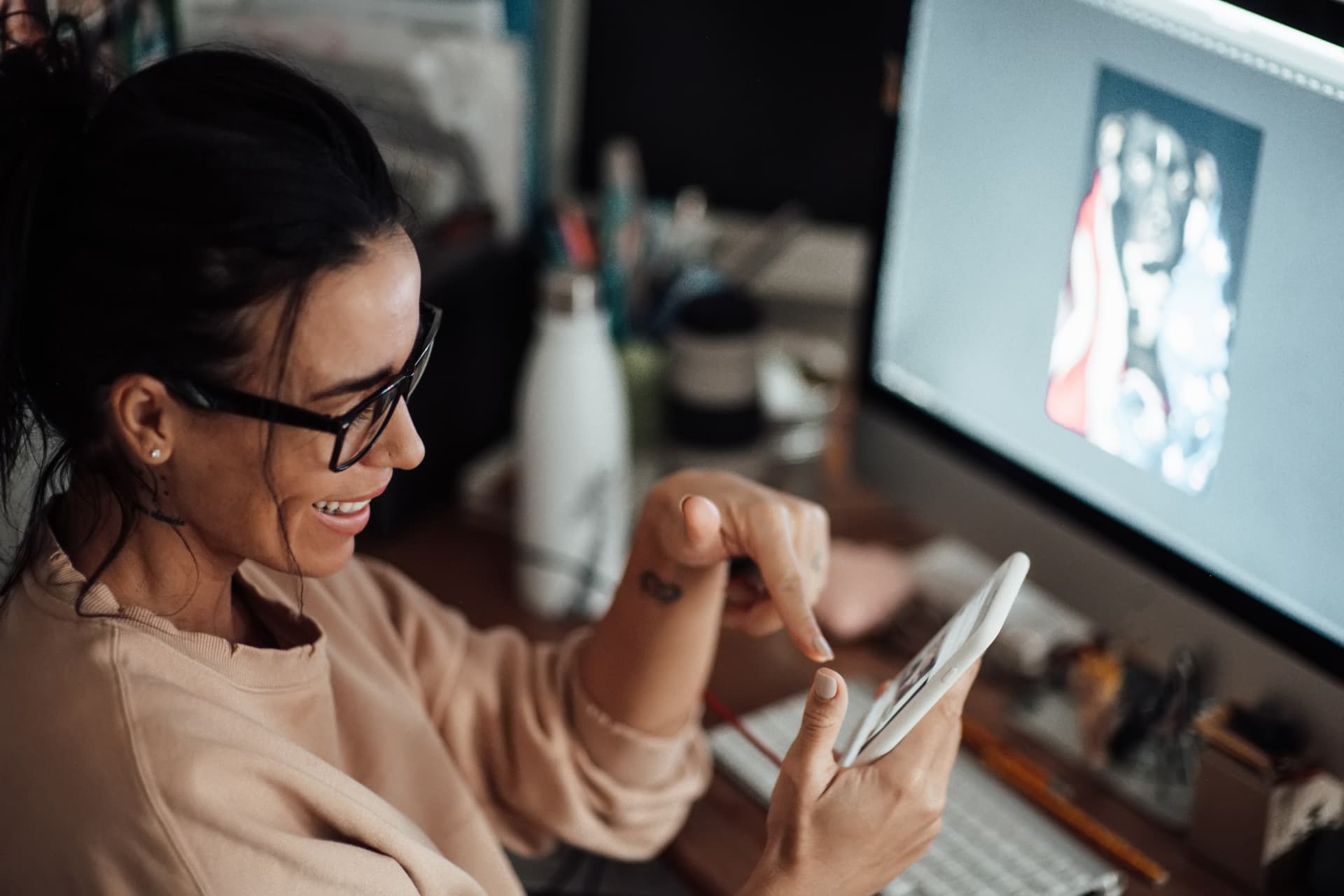 Mulher usando um smartphone enquanto trabalha diante de um monitor, ilustrando a produção criativa digital