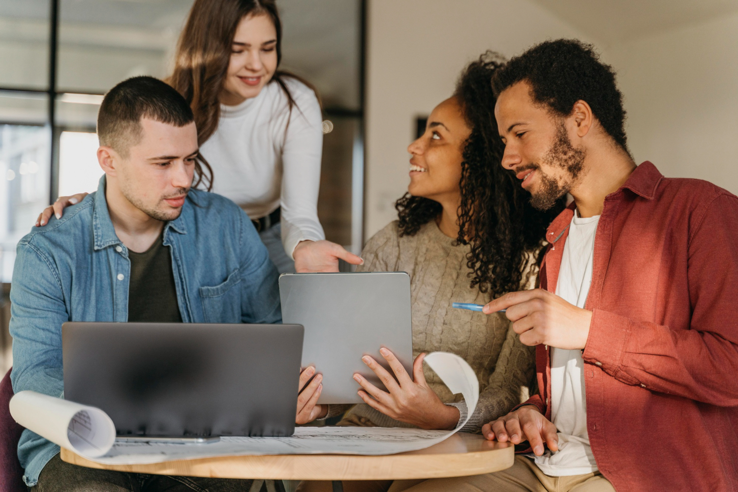 Equipe de marketing de uma agência reunida em volta de uma mesa usando ferramentas de produtividade em laptop e tablet para organizar as entregas dos clientes